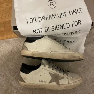Golden Goose vita sneakers med stjärna - Golden Goose sneakers i vitt skinn med grå mockadetaljer och ikonisk stjärna på sidan. Grå snörning, svart häl och beige sula ger en cool, avslappnad vibe. Perfekta för dig som gillar streetstyle och vill sticka ut med en unik look.