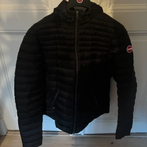 Svart dunjacka från Colmar - Snygg svart dunjacka från Colmar med horisontella sömmar och huva. Jackan har dragkedja framtill, två fickor med dragkedja och Colmar-logga på ärmen. Knappt använd 