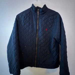 Quiltad marinblå jacka Polo Ralph Lauren - Säljer en quiltad marinblå jacka från Polo Ralph Lauren med dragkedja och två fickor framtill. Jackan har diskret röd logga på bröstet och snygga detaljer som tryckknappar. Perfekt för dig som gillar klassisk och stilren look.