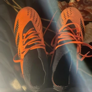 Adidas Predator orange fotbollsskor - Säljer ett par Adidas Predator fotbollsskor i orange och svart med coola detaljer och snörning. Skorna har hög strumpliknande krage och mönstrad ovandel för extra grepp på planen. Perfekta för dig som vill sticka ut på matchen!