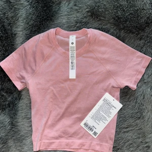 Rosa croppad t-shirt från Lululemon - Snygg croppad t-shirt från Lululemon i ljusrosa nyans. Fick av mina föräldrar när de var i usa och kunde tyvärr inte lämna tillbaka🫶🏼 ALDRIG ANVÄND!!T-shirten har korta ärmar, rund halsringning och diskret randigt mönster. Perfekt för träning eller chill dagar. Materialet är mjukt och andas, vilket gör den superbekväm.