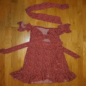 Röd blommig omlottklänning med volang - Supersöt röd kortklänning med vitt blommönster, omlottmodell och volangkant nertill. klänningen går att knyta på flera olika sätt, ett kortare band (se bild) tillkommer. Klänningen har knytband i midjan, korta ärmar med volang och en snygg rosettdetalj framtill. 