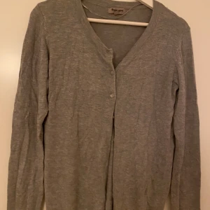 Grå somrig kofta - En enkel och stilren grå kofta från Basic You i storlek S. Koftan har v-ringning, knäppning framtill med små knappar och långa ärmar. Perfekt att slänga över en t-shirt eller linne.