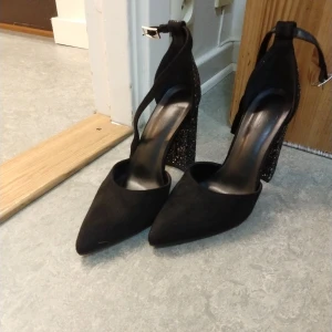 Svarta pumps med glittrig klack - Snygga svarta pumps med spetsig tå och bred, hög klack täckt av svart glitter. Skorna har rem runt vristen med spänne och är gjorda i mockaliknande material. Perfekta för dig som vill sticka ut med en cool och glamorös look.