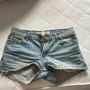 Ljusblå jeansshorts från Levi's - Säljer ett par ljusblå jeansshorts från Levi's med råa, fransiga kanter och slitna detaljer. Klassisk femficksmodell med knapp och dragkedja framtill. Perfekta för varma dagar.