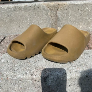 Yeezy Slide 'Ochre' sandaler - Säljer ett par Yeezy Slide 'Ochre' från Adidas i en beige-brun färg. Sandalerna har öppen tå och platt sula med en chunky design. Perfekta för sommaren och riktigt bekväma att glida runt i. Skorna är köpta på pardonmykicks i Malmö när det fanns där. Kvitto finns! Inga direkta defekter förutom normalt slitage. Om det är något mer du undrar är det bara att skriva😃