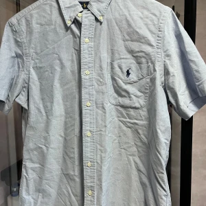 Ljusblå kortärmad skjorta från Ralph Lauren - Säljer en klassisk ljusblå kortärmad skjorta från Ralph Lauren med button-down krage och bröstficka med broderad logga. Skjortan har knappar framtill och är tillverkad i bomull. Perfekt för en stilren och avslappnad look.