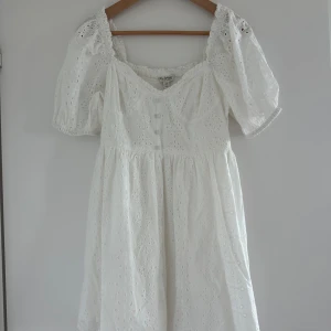 Vit klänning med puffärmar och broderade detaljer - Superfin vit klänning med broderade hålmönster, puffiga kortärmar och v-ringning framtill. Dekorativa knappar framtill och smock i ryggen för extra komfort. Perfekt för sommarens alla tillfällen! Aldrig använd.