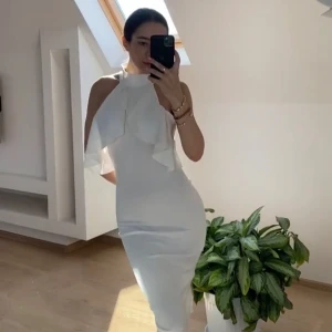 Vit halterneckklänning med volangdetalj - Säljer en elegant vit halterneckklänning med volang framtill och hög hals. Klänningen är figurnära och har en stilren, modern look som passar perfekt till speciella tillfällen.