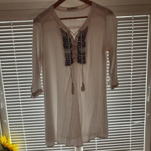 Vit blus med knytning från Mango - Luftig vit blus från Mango med trekvartsärm och dekorativ knytning framtill. Svart broderad detalj vid bröstet och tofsar i snörena. Perfekt för varmare dagar eller som strandplagg.