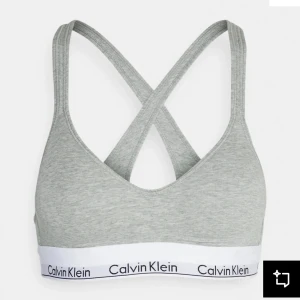 Grå sport-bh från Calvin Klein - Sport-bh i ljusgrå bomull med bred resår och Calvin Klein-logga i vitt och svart. Korsade axelband i ryggen för extra stöd och snygg look. Perfekt för träning eller chill dagar.