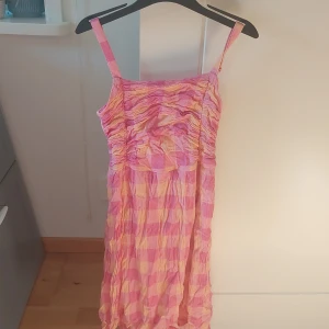Rosa och gul rutmönstrad klänning med smock - Säljer en söt klänning med rutigt mönster i rosa och gul. Klänningen har smala axelband, smockad överdel och lätt rynkad kjol. Perfekt för varma dagar och har en luftig känsla.