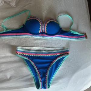 Blå bikini med färgglada detaljer  - Säljer en snygg blå bikini med vadderad bygel-bh och matchande trosa. Båda delarna har dekorativa sömmar i rosa, grön och vit längs kanterna. Axelbanden är mintgröna och bikinin har en sportig och färgglad look. Använt skick men inga fläckar eller liknande