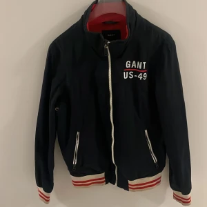 Blå vindjacka från Gant med tryck - Säljer en blå vindjacka från Gant med vitt tryck 'GANT US-49' på bröstet och koordinater på ryggen. Jackan har dragkedja, vita och röda ribbade muddar samt två fickor med dragkedja. Insidan är röd med meshfoder. Pris kan diskuteras!