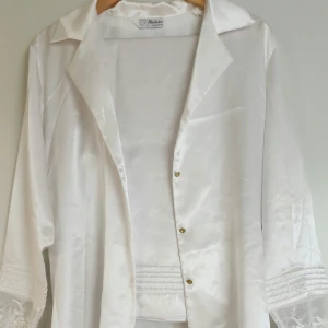 Tvådelat pyjamas-set i vit satin - Elegant och bekvämt set med skjortöverdel och byxor. Spetsdetaljer på ärmslut, guldknappar och mjukt, glansigt tyg.                                                Perfekt för dig som vill känna dig både bekväm och fin hemma eller vid speciella tillfällen.    