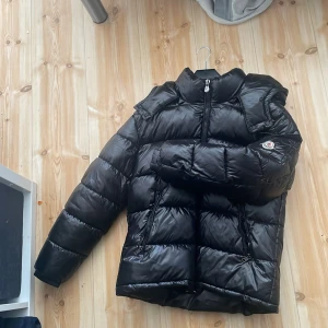 Svart pufferjacka från Moncler - Säljer en svart pufferjacka från Moncler med hög krage och dragkedja. Jackan har Moncler-logga på ärmen och klassisk quiltad design. Perfekt för kyliga dagar och riktigt snygg till streetwear-stilen.