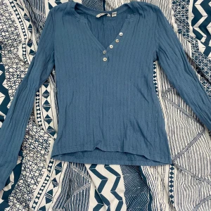 Blå ribbad långärmad tröja från Aware - Säljer en blå ribbad långärmad tröja från Aware i storlek XS. Tröjan har v-ringning med dekorativa knappar framtill och är tillverkad i ett mjukt material. Knappt använd. 150 kr