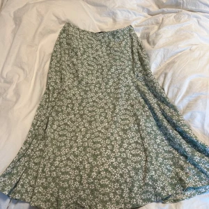 Grön blommig långkjol från DAZY - Säljer en grön långkjol från DAZY med vitt blommönster. Kjolen har en lätt utställd form och en diskret slits på sidan för extra rörelsefrihet. Perfekt för dig som gillar en feminin och avslappnad stil.