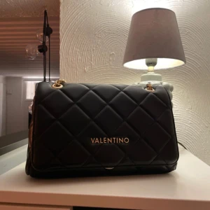 Svart quiltad axelväska från Valentino - Säljer en svart quiltad axelväska från Valentino med guldfärgade kedjedetaljer och logotyp framtill. Väskan har ett elegant rutmönster och stängs med dragkedja. Perfekt för dig som vill ha en stilren accessoar med lyxig känsla.