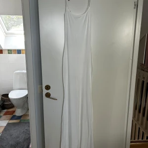 Vit långklänning med smala axelband - Äkta Skims klänning. En enkel och stilren vit långklänning med smala axelband. Klänningen har rak passform och är tillverkad i ett mjukt, stretchigt material som ger en bekväm känsla. Perfekt för varma dagar eller sommarens alla tillfällen.