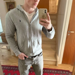 Grå hoodie med dragkedja från Polo Ralph Lauren - Säljer en stilren grå hoodie från Polo Ralph Lauren med dragkedja och huva. Jackan har två fickor framtill och en broderad logga på bröstet. Perfekt för en avslappnad look.