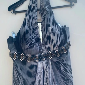 Grå långklänning med djurmönster st.34-38 och pärldetaljer - Säljer en snygg grå långklänning med djurmönster och halterneck.passar 34-38(40). Klänningen har markerad midja med pärl- och stendekorationer samt djup v-ringning. Tyget är lätt och faller fint, perfekt för fest eller bal.