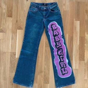 Blå jeans med tryck  - Unika blå jeans med stort airbrush-tryck i lila och rosa med texten 'BABY GIRL' längs ena benet. Kontrast-sömmar i rosa och hjärtan på bakfickorna. Rak passform och klassisk femficksmodell. Perfekt statement-plagg för dig som vill sticka ut.