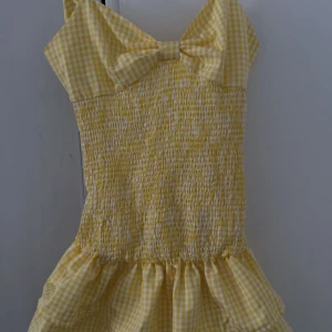 Gul rutig klänning med rosett - Supersöt gul och vit rutig klänning med smockad överdel och stor rosett framtill. Klänningen har smala axelband och volang nertill, perfekt för soliga dagar. Lätt och luftig modell med knyt i ryggen. Strl XS och är perfekt till sommaren 💕