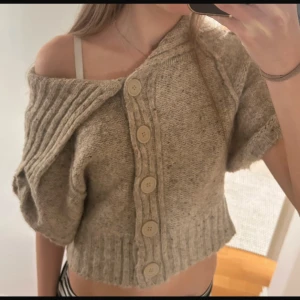 Beige stickad croppad kofta med stora knappar - Säljer en beige stickad kofta med korta ärmar och stora knappar framtill. Koftan är croppad och har en bred ribbad krage som kan bäras off-shoulder. 