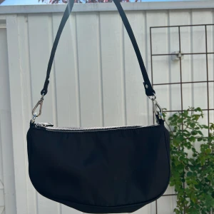 Svart axelväska med silverfärgad dragkedja - Snygg svart axelväska med stilren design och silverfärgad dragkedja upptill. Väskan har en avtagbar axelrem och passar perfekt för att bära det viktigaste. Enkel och trendig modell som funkar till många outfits.