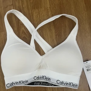Vit sport-bh från Calvin Klein - Säljer en vit sport-bh från Calvin Klein med korsade axelband och elastiskt band med logga nedtill. Gjord i mjuk bomullsmix, perfekt för träning eller vardag. Modern Cotton-kollektionen ger en stilren och bekväm känsla.