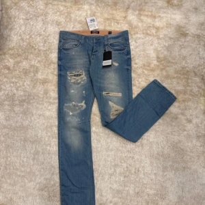 Ljusblå slitna jeansbyxor från Sugar Cane - Ljusblå jeansbyxor från Sugar Cane med flera slitningar och lagade detaljer framtill. Klassisk femficksmodell med normal passform och raka ben. Jeansen har en vintagekänsla och är perfekta för en avslappnad stil.