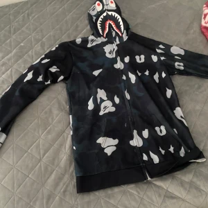 Svart  hoodie med hajmotiv  - Säljer en svart  hoodie  med hajmotiv på huvan och hel dragkedja. Jackan har unikt mönster i grått och vitt samt fickor framtill. Perfekt för dig som gillar streetwear och vill sticka ut.