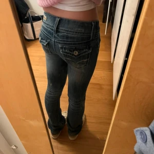 Blå lågmidjade jeans från Refuge - Säljer ett par blå lågmidjade jeans från Refuge med slitningar och dekorativa bakfickor med knappar och vita sömmar. Jeansen har en klassisk straight fit och är perfekta för dig som gillar en avslappnad stil. Innerbenslängden är 78cm och midjemåttet är ca 34cm💓