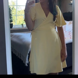 Ljusgul omlottklänning med kort ärm - Söt ljusgul omlottklänning med v-ringning och knyt i midjan. Klänningen har korta, vida ärmar och en lätt utsvängd kjol som slutar ovanför knäna. Perfekt för varma dagar och somriga tillfällen.💞