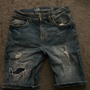 Blå jeansshorts från LAB Industries - Säljer ett par blå jeansshorts från LAB Industries med slitna detaljer och råa kanter. Klassisk femficksmodell med knapp och dragkedja i gylfen.