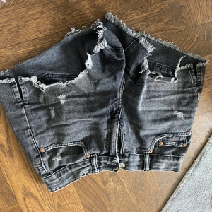 Svarta jeansshorts med fransar från Zara - Säljer ett par svarta jeansshorts från Zara med fransiga kanter och slitna detaljer. Shortsen har resår i midjan och klassiska fickor både fram och bak. Perfekta för en avslappnad stil.