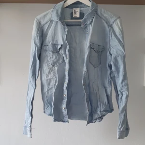Ljusblå jeansskjorta från H&M - Ljusblå jeansskjorta från H&M med tryckknappar och två bröstfickor med lock. Skjortan har lång ärm och klassisk krage. Perfekt för en avslappnad och stilren look.