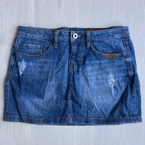 Blå jeanskjol från edc - Säljer en klassisk blå jeanskjol från edc med femficksmodell och synliga slitningar framtill. Kjolen har bälteshällor och knappstängning i midjan. Perfekt till sneakers eller boots.