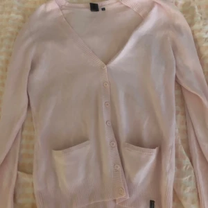 Ljusrosa kofta från Global Sport - Säljer en ljusrosa kofta från Global Sport i storlek M. Koftan har v-ringning, knäppning framtill med flera knappar och två fickor framtill. Perfekt för lager-på-lager och enkel att matcha med olika outfits.