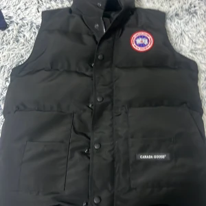 Svart dunväst från Canada Goose - Säljer en svart dunväst från Canada Goose med klassisk logotyp på bröstet och diskret varumärkesmärke på fickan. Västen har hög krage, knappar och dragkedja framtill samt två stora fickor.
