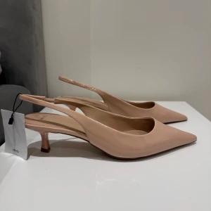 Klackar/pumps/sandaler från Mango - Säljer ett par stilrena beige pumps från Mango med spetsig tå och låg klack. Skorna har öppen häl med smal rem och är tillverkade i glansigt syntetmaterial. Perfekta för dig som gillar en klassisk och elegant look. NYPRIS 420kr!! Storlek 40, helt slutsålda!! 🥰
