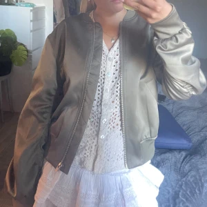 Olivgrön bomberjacka - Snygg olivgrön bomberjacka med dragkedja framtill och ribbade muddar vid ärmslut och midja. Jackan har en klassisk bombermodell och är tillverkad i ett slätt, glansigt material. 