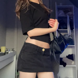 Svart lågmidjad jeanskjol med knapp - Säljer en svart kort jeanskjol med klassisk knapp och fickor framtill. Kjolen har en rak passform och är perfekt för dig som gillar en enkel och stilren look.