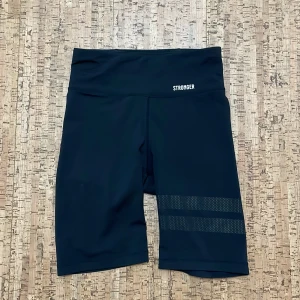 Svarta cykelshorts från Stronger - Svarta cykelshorts från Stronger med hög midja och diskreta randiga detaljer på ena benet. Tillverkade i stretchigt syntetmaterial som passar perfekt för träning. Logotypen är tryckt framtill i vitt.