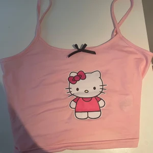 Rosa Hello Kitty linne - Säljer ett ljusrosa linne med smala axelband och en söt Hello Kitty-tryck framtill. Toppen har en liten svart rosett vid halsringningen och är croppad i modellen. Använd 1 gång