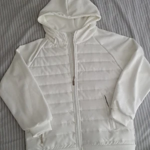 Vit Cardigan  - Snygg vit hoodie med quiltade paneler och dragkedja framtill. Jackan har huva, två fickor med dragkedja och ribbade muddar vid ärmslut och nederkant. Perfekt för en stilren och sportig look.                                                                           Den är ny och oanvänd.