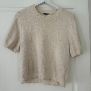 Beige stickad kortärmad tröja - Säljer en beige stickad tröja med korta ärmar och rund halsringning. Tröjan har en enkel och stilren design med ribbade muddar vid ärmslut och nederkant. Perfekt för lager-på-lager eller sommarens svalare dagar.