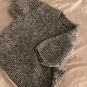 Grå stickad tröja från Gina Tricot - Mysig grå stickad tröja från Gina Tricot med korta ärmar och rund halsringning. Tröjan har en klassisk, enkel design och är perfekt för lager-på-lager-stil under kyligare dagar.