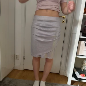 Silverfärgad glittrig omlottkjol - Säljer en snygg silverfärgad kjol med glittrigt tyg och omlottdetalj framtill. Kjolen är figurnära och har en kort längd som passar perfekt till festliga tillfällen. Enkel att matcha med olika toppar för en trendig look.